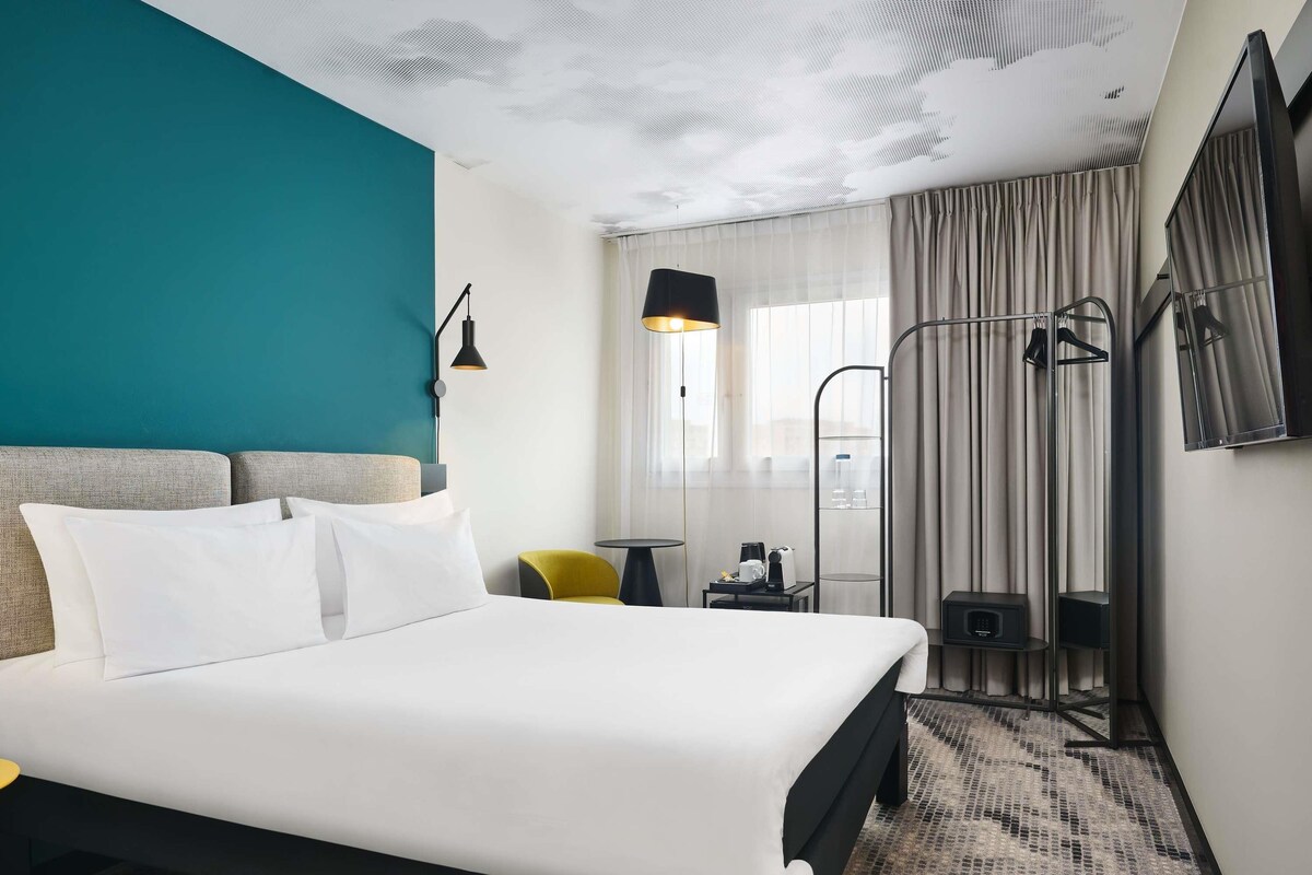 A hotel room with a bed, a desk, a chair, a TV, and a window with curtains.