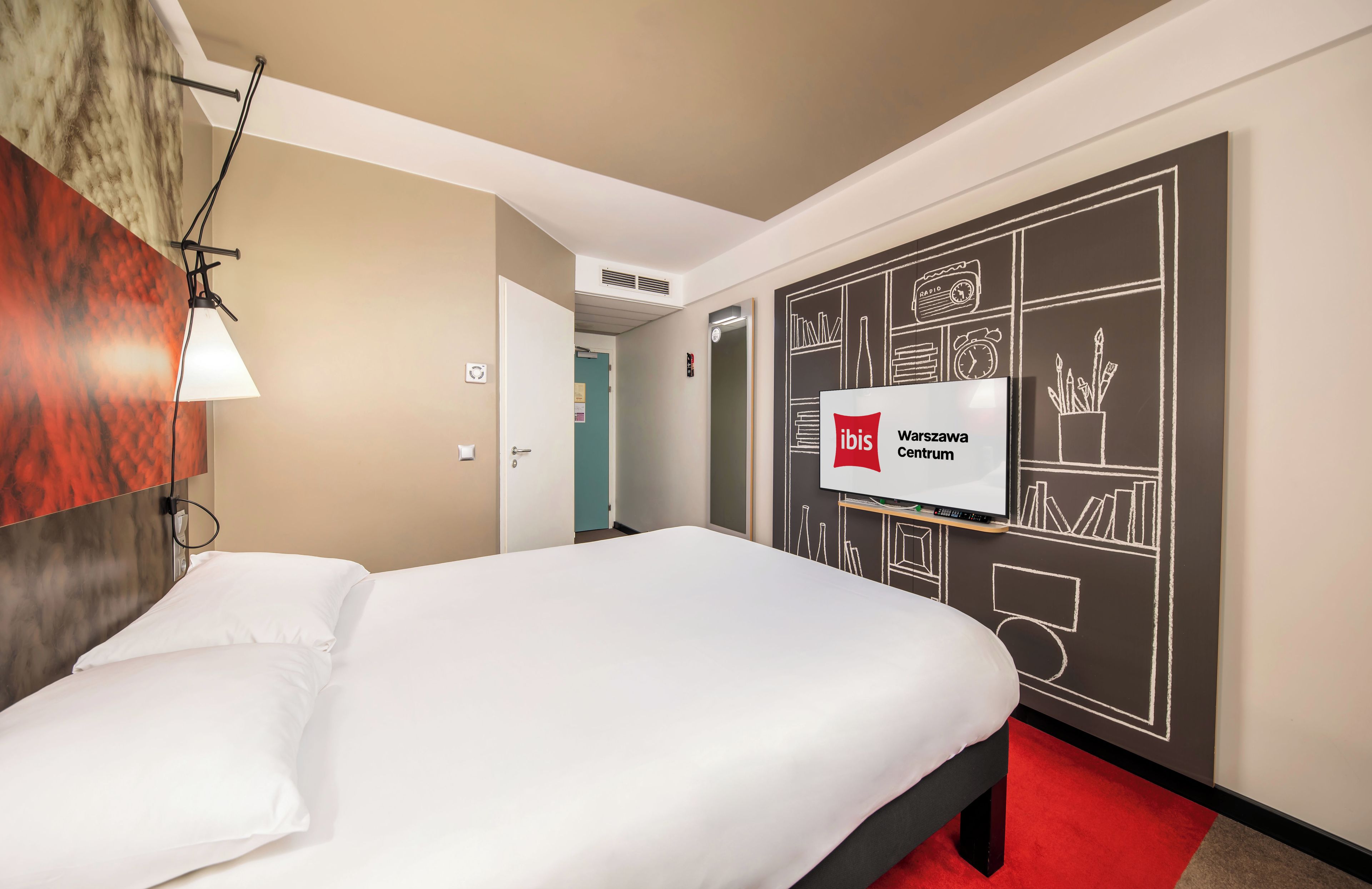 A hotel room with a bed, a wall-mounted lamp, a sign indicating 'ibis Warszawa Centrum', and a chalkboard wall with drawings.
