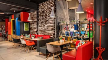 Um café moderno com assentos vermelhos e pretos, mesas de madeira e uma variedade de plantas e livros nas prateleiras.