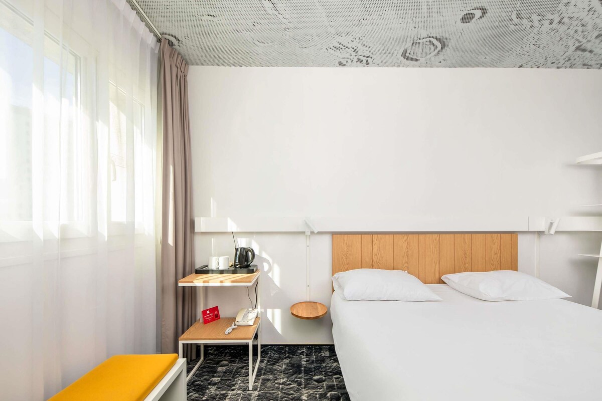 A hotel room with a bed, a bedside table with a lamp, a small table with a kettle and cups, a bench with a yellow cushion, and a patterned carpet.