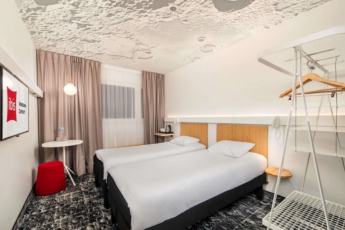A hotel room with a double bed, a small table with a coffee maker, a red chair, and a ceiling with a unique pattern.