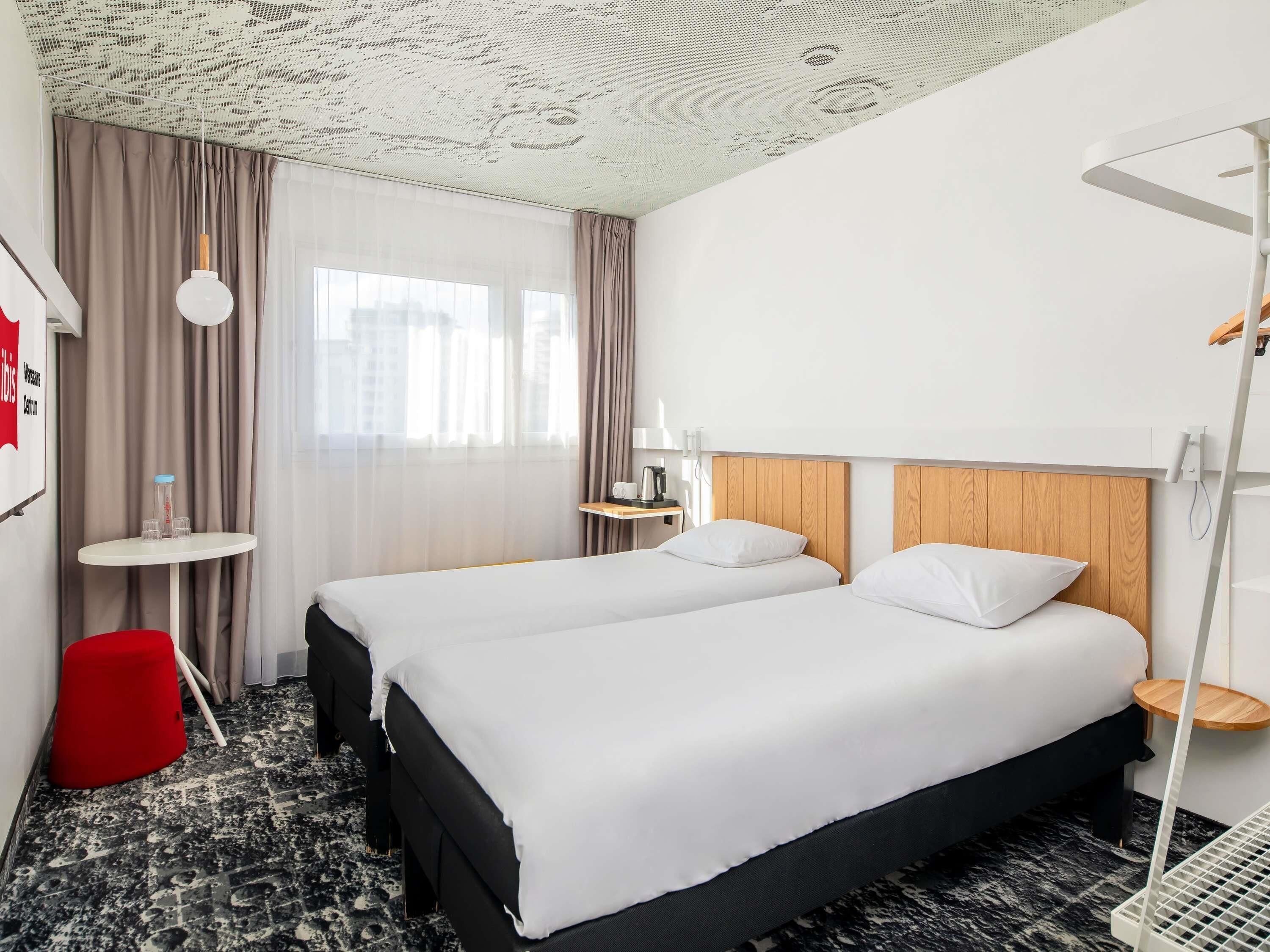 Une chambre d’hôtel avec deux lits, une petite table, un tabouret rouge et un plafond orné d’un motif original.