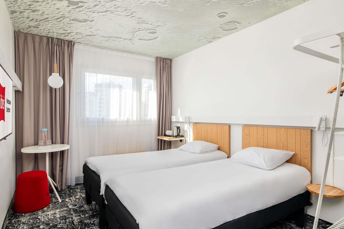 A hotel room with two beds, a red stool, a small table, and a ceiling with a unique pattern.