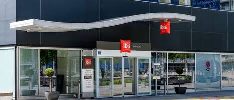 Un edificio moderno con entrada de vidrio, el letrero rojo de 'ibis' y una rampa accesible para sillas de ruedas.