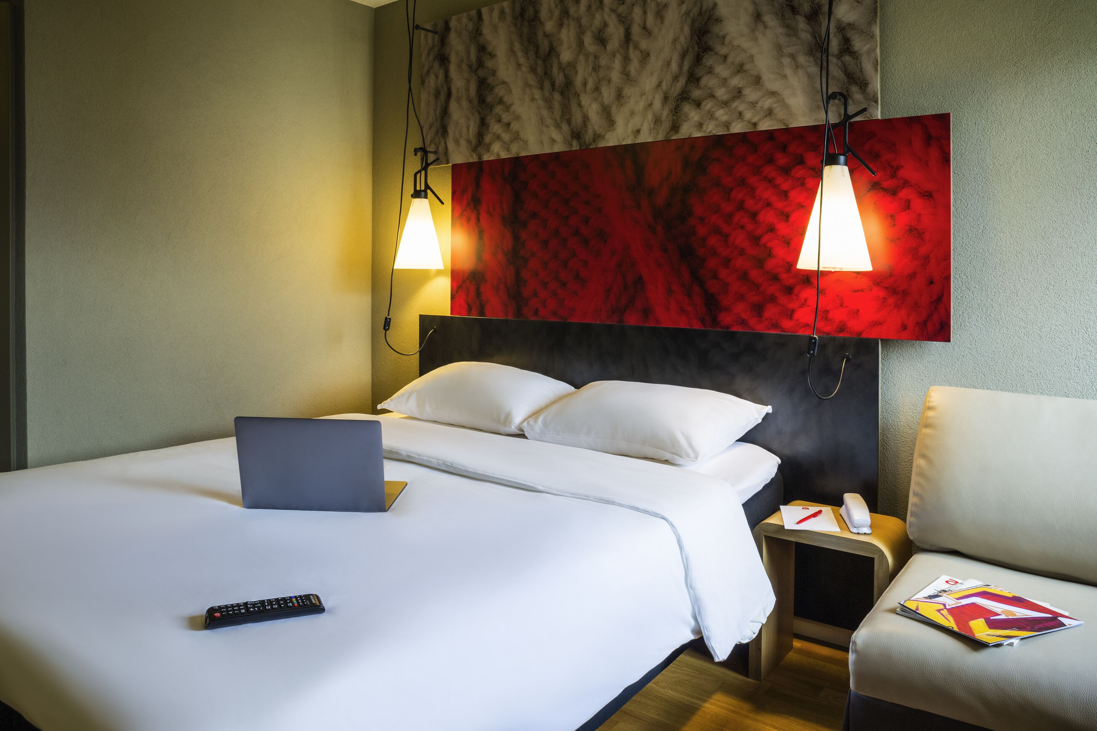 A hotel room with a bed, a laptop, a remote control, a nightstand with a phone and a magazine, and two pendant lights.