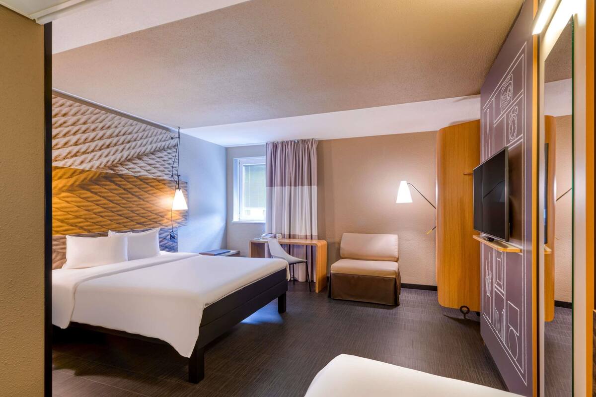 Ein Hotelzimmer mit einem Bett, einem Schreibtisch, einem Sessel, einem Fernseher und einem Fenster mit Vorhängen.