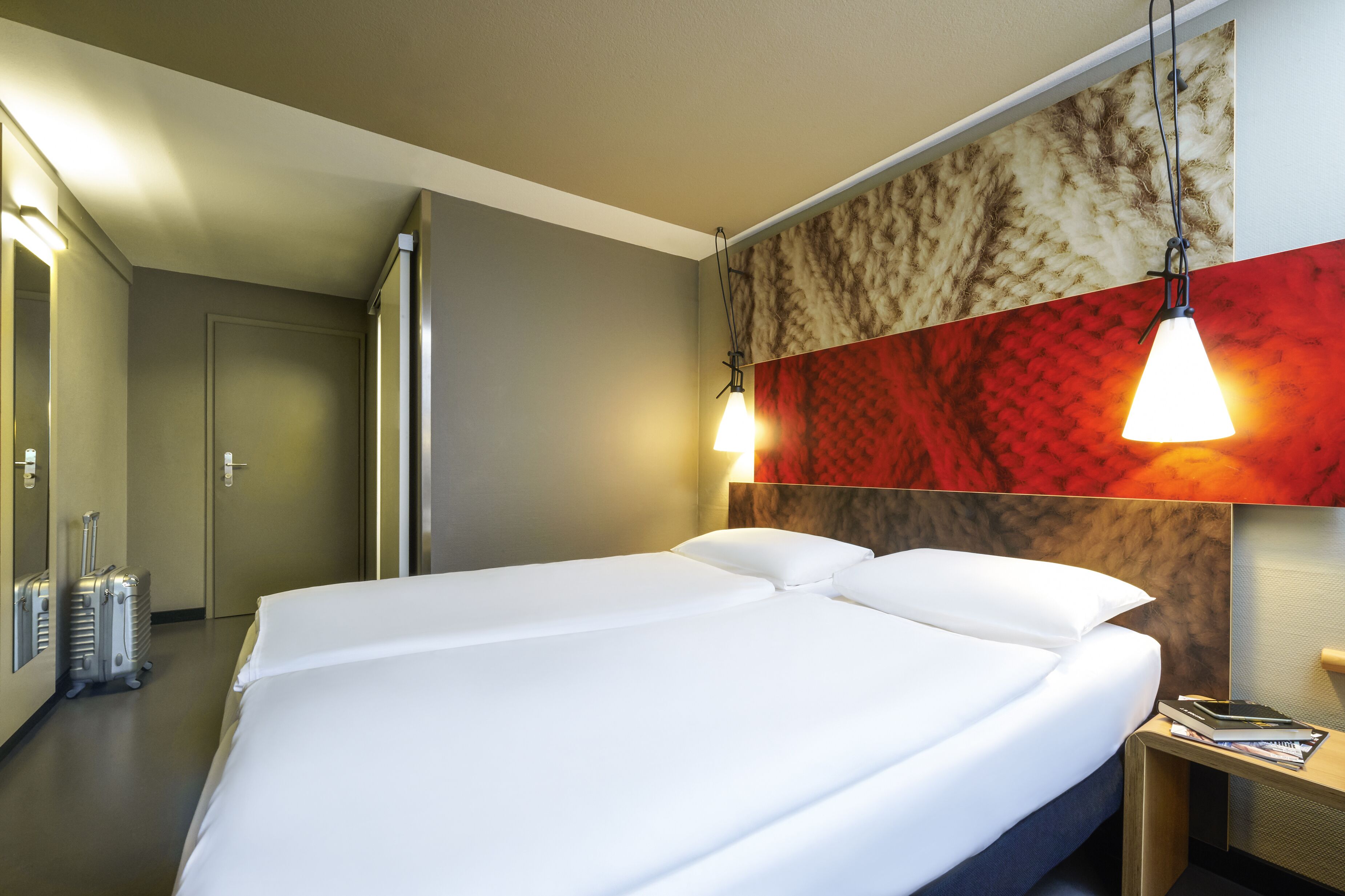 A hotel room with a large bed, a nightstand with a lamp, a suitcase, and a door to another room.