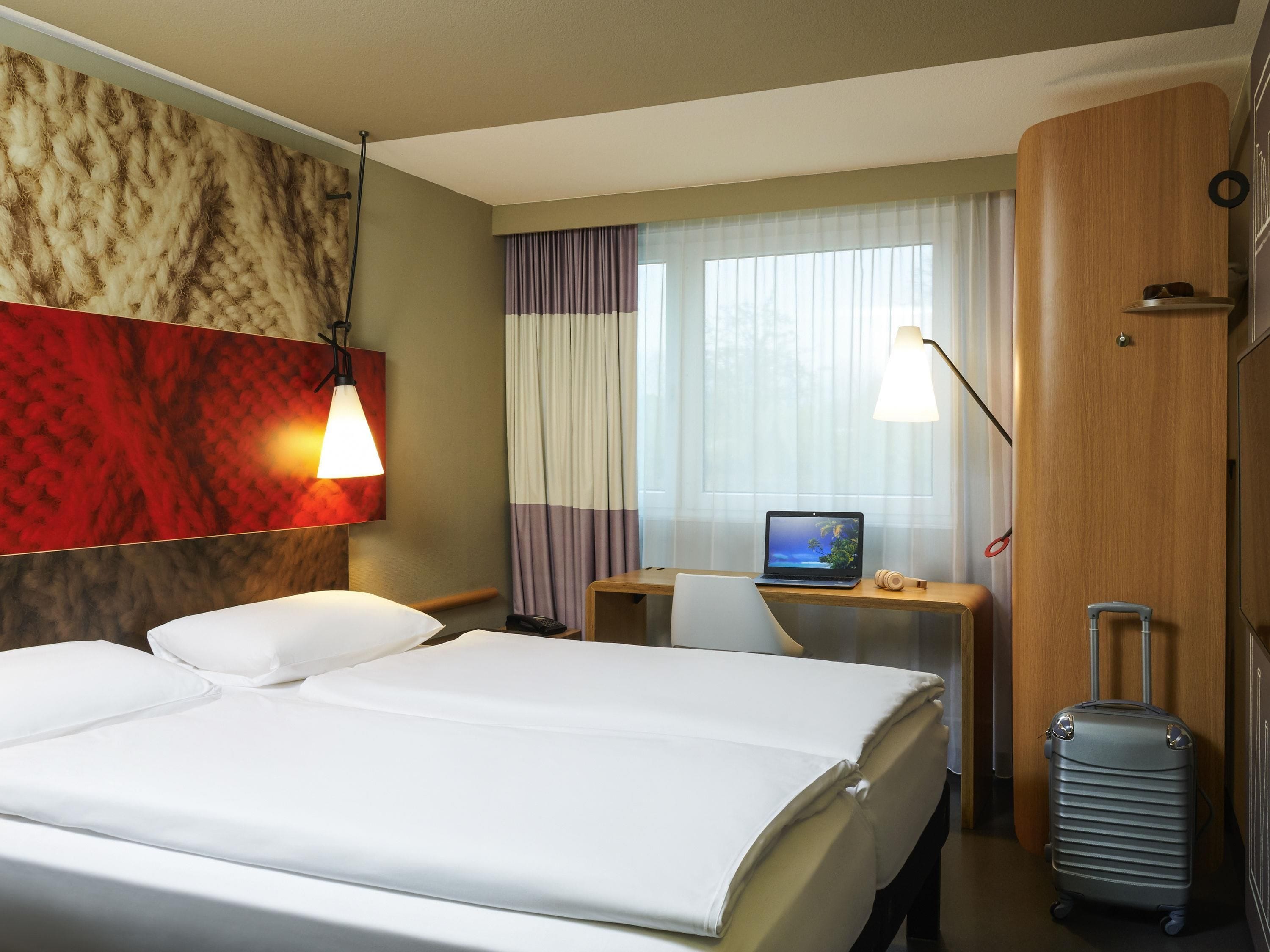 A hotel room with a bed, a desk with a laptop, a suitcase, and a lamp.