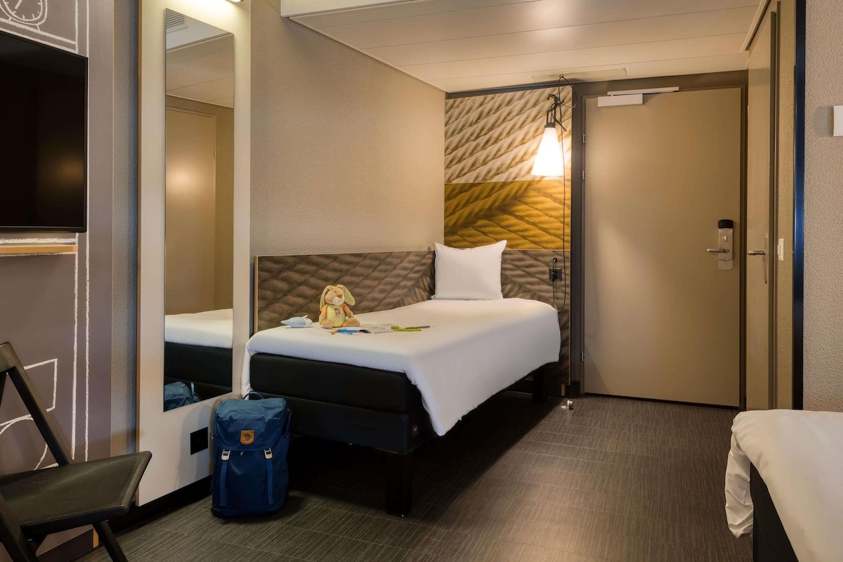 A hotel room with a bed, a chair, a desk, a suitcase, and a mirror.