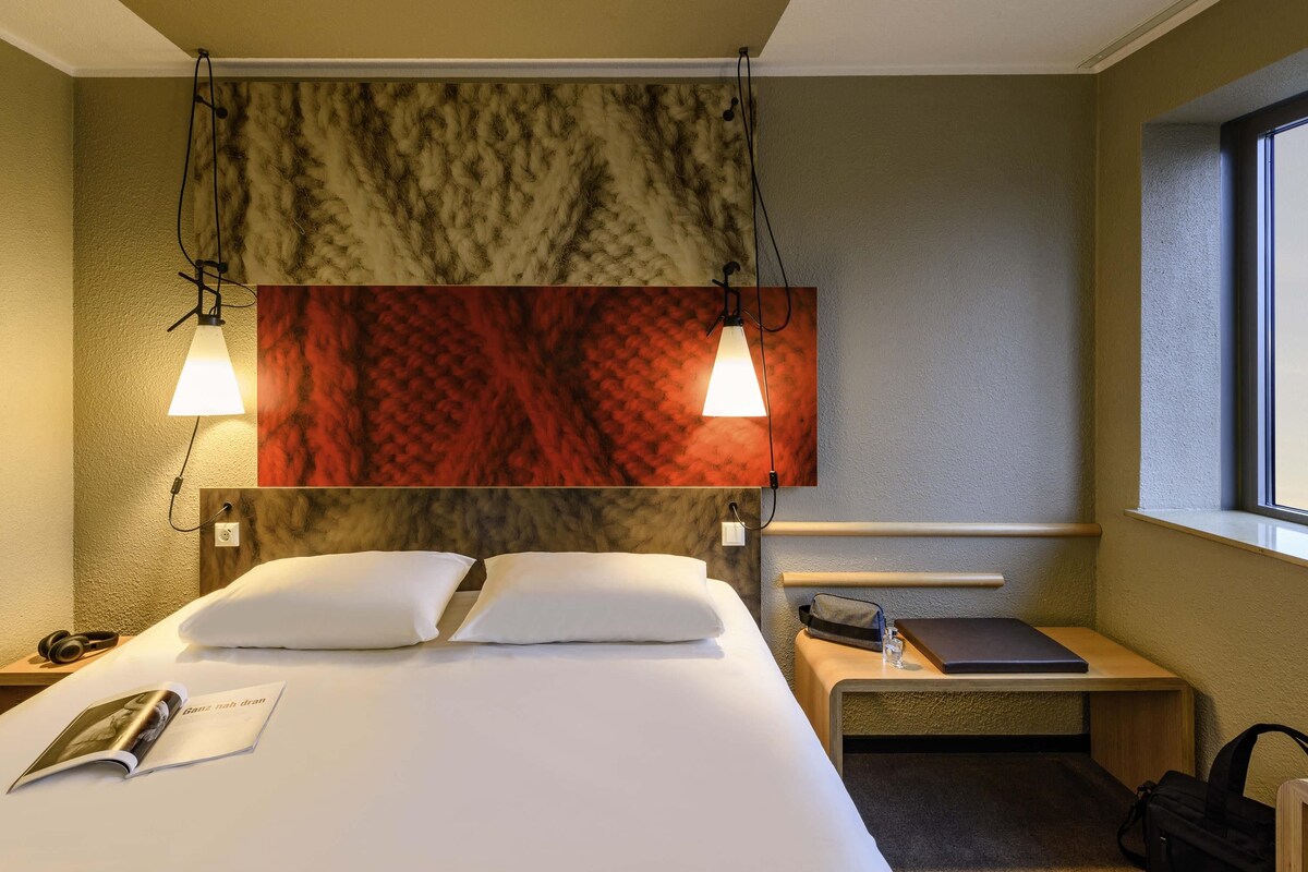 A hotel room with a bed, bedside tables, a nightstand, a reading lamp, a window, and a wall-mounted artwork.