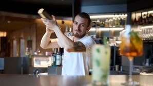 Un barman prépare un cocktail derrière un comptoir, avec diverses bouteilles et différents verres en arrière-plan.