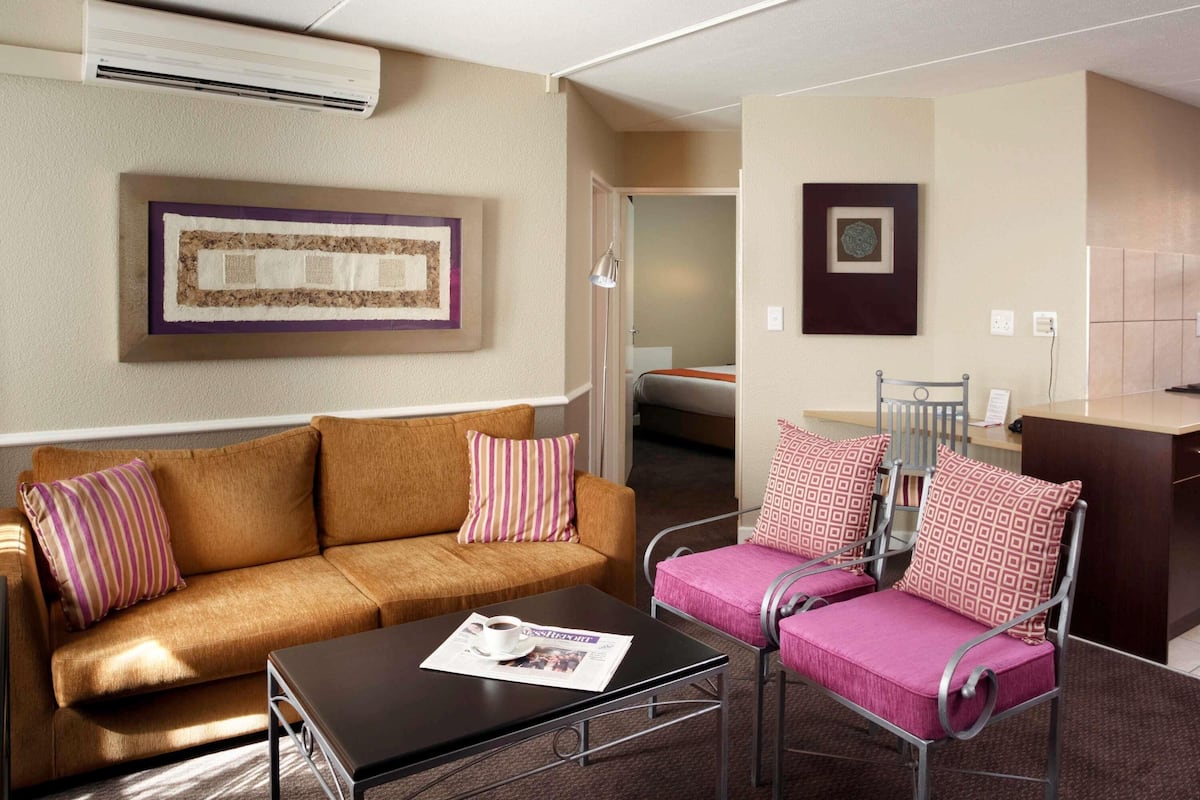 A hotel room with a brown sofa, a small table, a metal chair with a pink cushion, a bed, a desk, and a wall-mounted air conditioner.