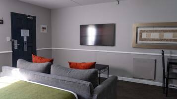 Habitación de hotel con cama, dos sillones, una mesita, un televisor de pantalla plana, un cuadro enmarcado y una puerta con un cartel.