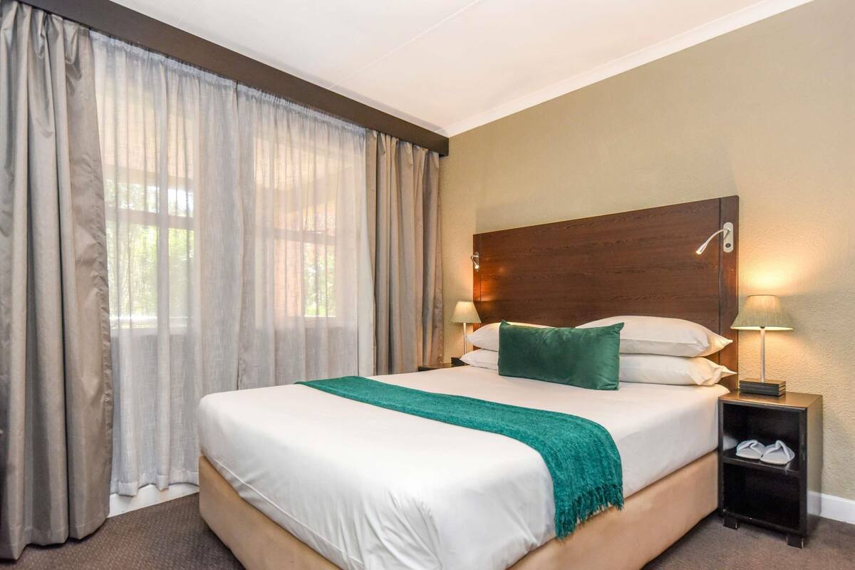 Hotellihuone, jossa on suuri sänky, puinen pääty, kaksi yöpöytävalaisinta, yöpöytä, jossa on kenkäteline, ja läpikuultavat verhot.