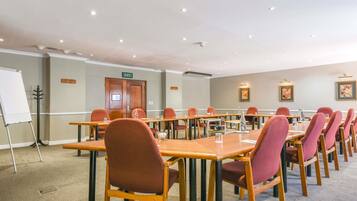 A conference room with multiple tables and chairs, an exit door, a whiteboard, and framed artwork on the walls.