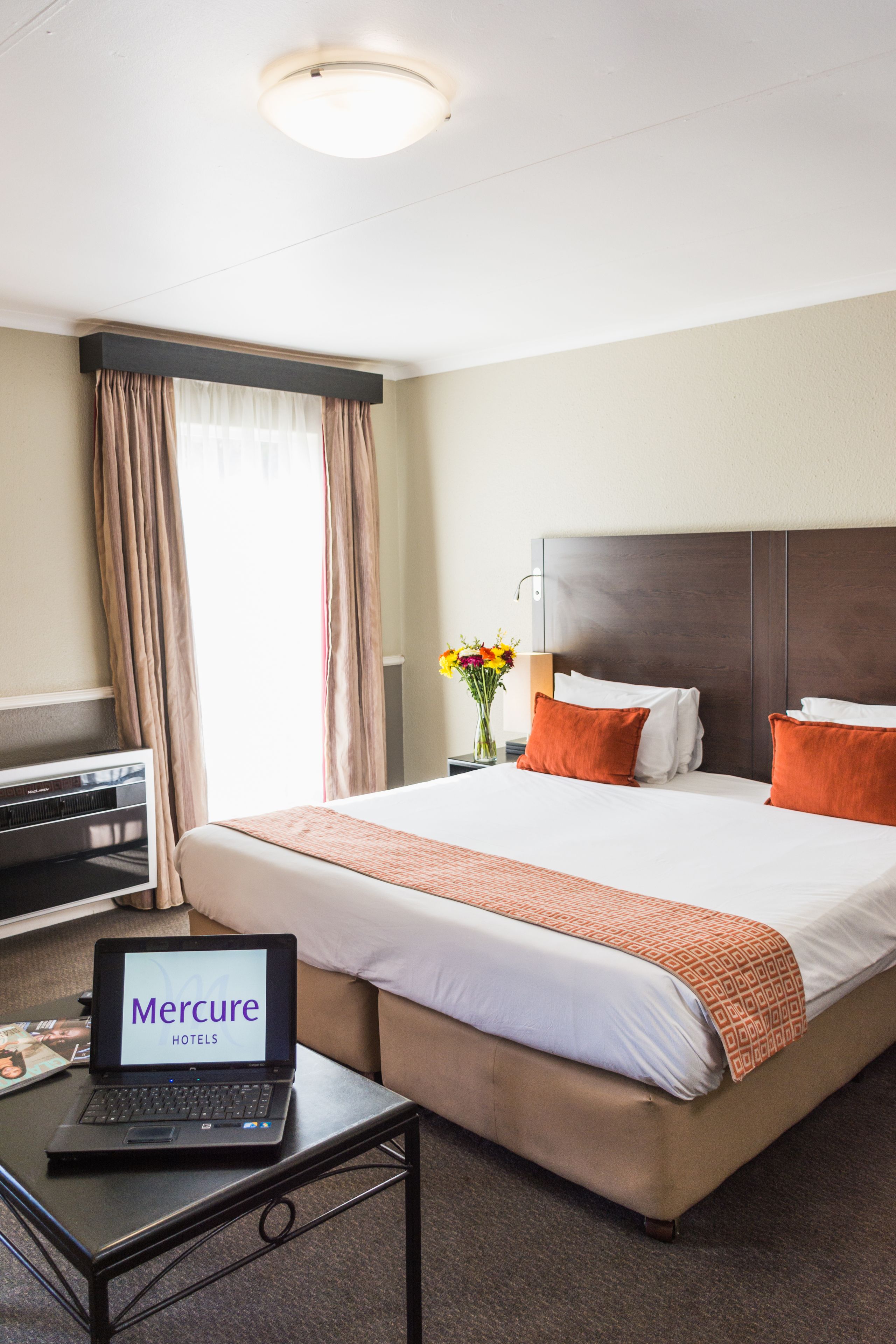 Ein Hotelzimmer mit Bett, Laptop auf einem Nachttisch, Schreibtisch und einem Fenster mit Vorhängen.