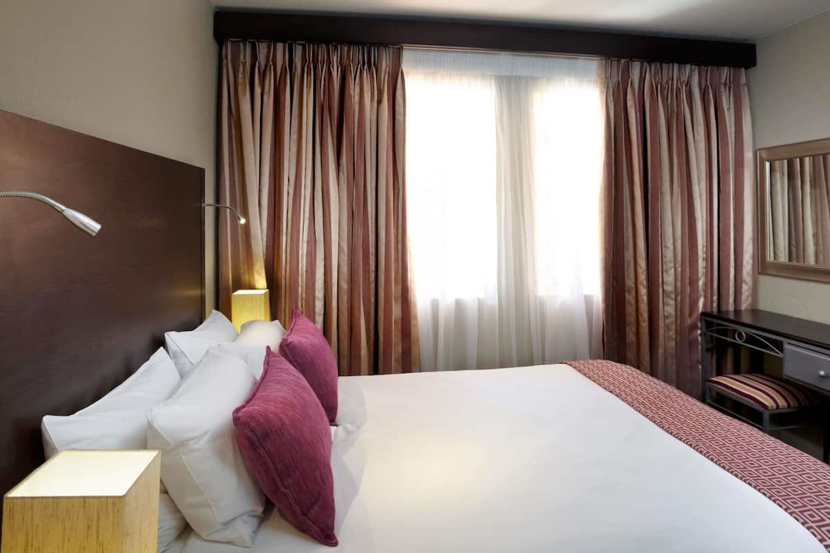 A hotel room with a large bed, a bedside table with a lamp, a desk with a chair, and a window with curtains.