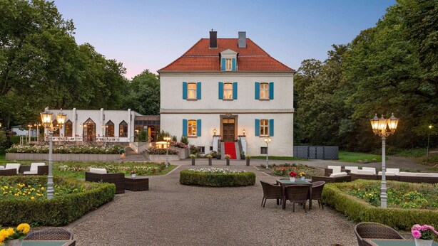 Un bâtiment à deux étages, surmonté d’un toit recouvert de tuiles rouges, entouré d’une cour en gravier aménagée avec des espaces de détente en plein air et des parterres de fleurs.