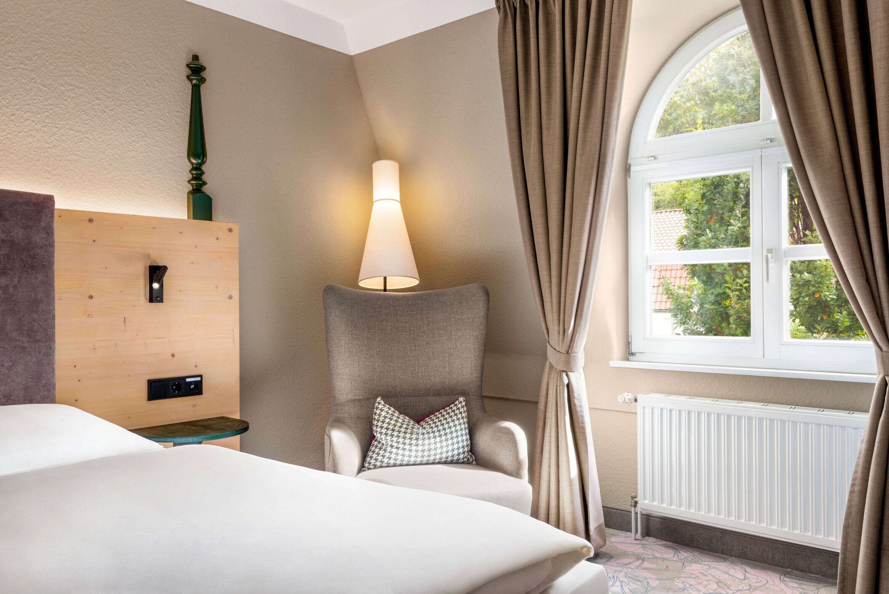 Habitación de hotel con cama, una silla con lámpara, pared de paneles de madera y ventana con cortinas.