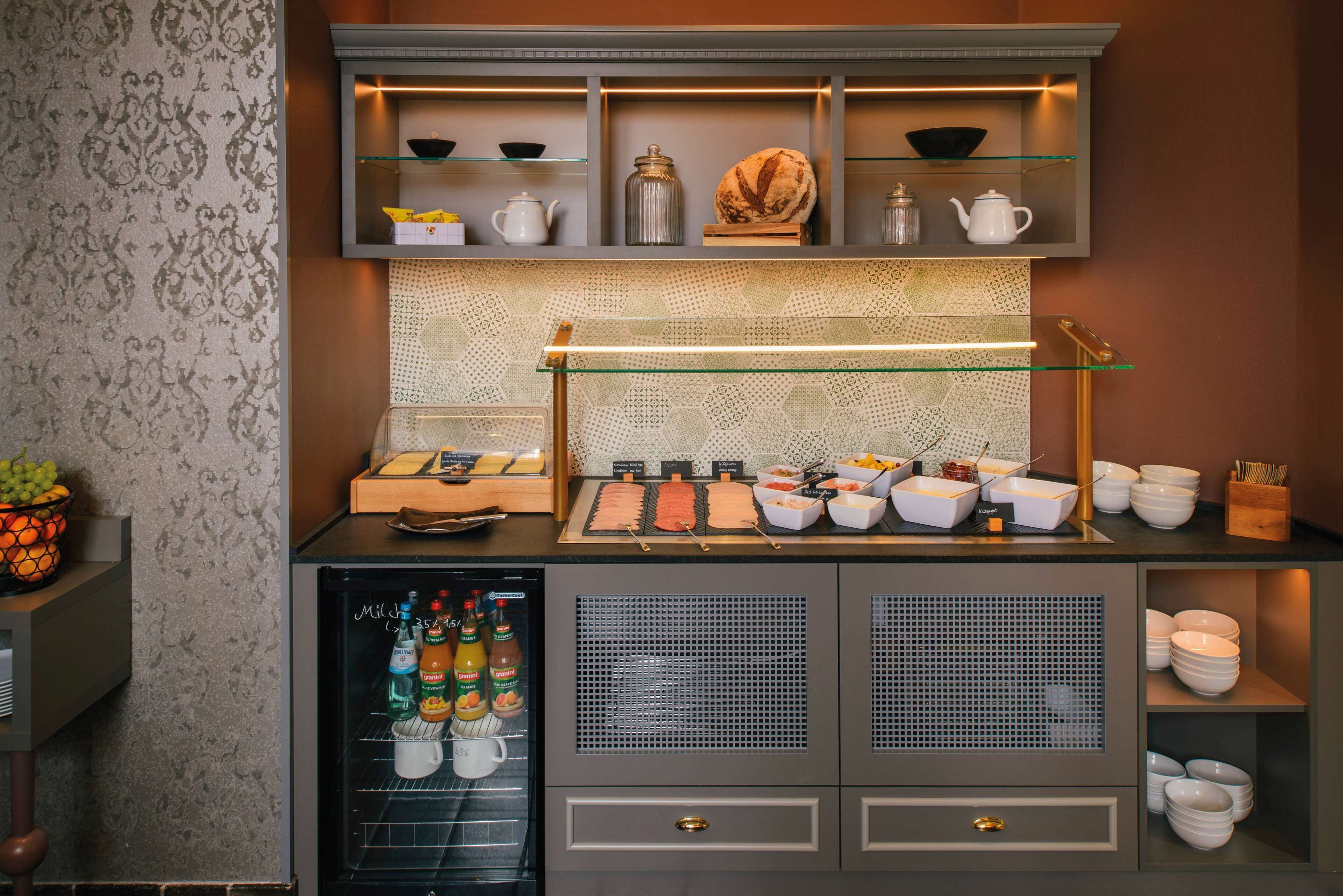 Una barra de desayuno con diversos alimentos y condimentos, un refrigerador con bebidas, y una pared decorativa con estantes.