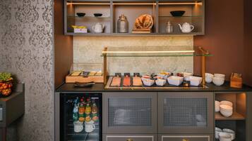 Una barra de desayuno con diversos alimentos y condimentos, un refrigerador con bebidas, y una pared decorativa con estantes.