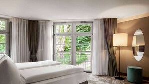 Une chambre d’hôtel avec un lit, une fenêtre avec des rideaux, une lampe de sol, un miroir et un pouf vert.