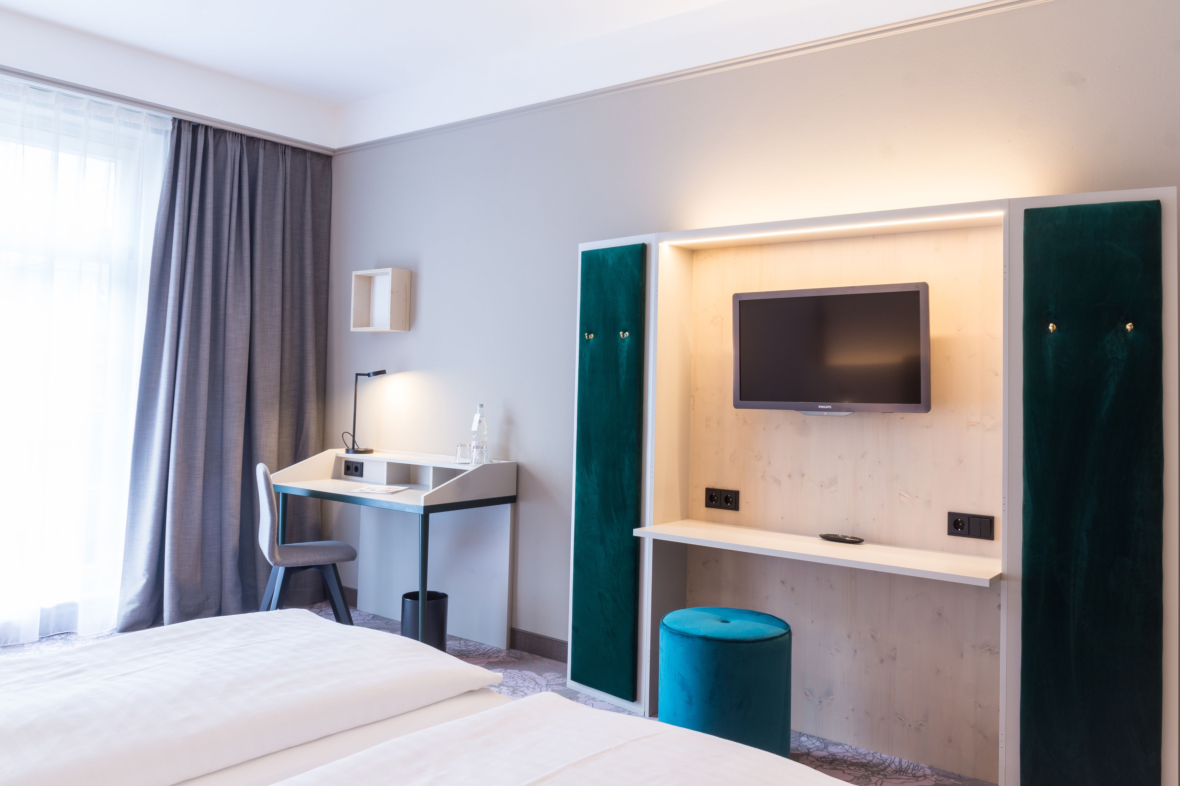 A hotel room with a bed, a desk with a chair, a TV mounted on the wall, and a window with curtains.