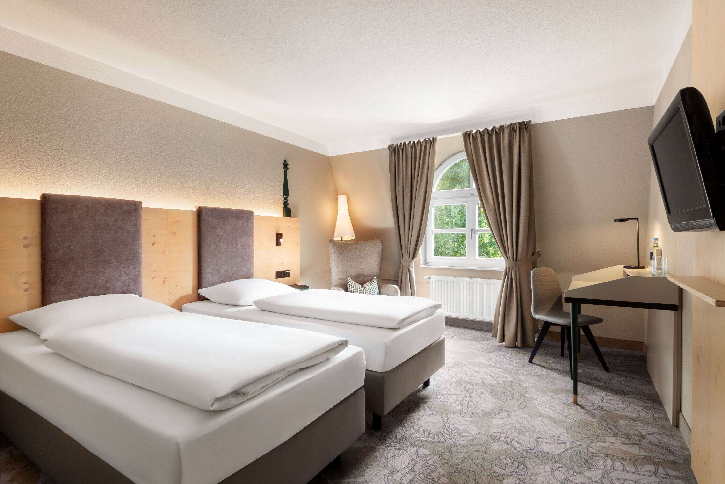 A hotel room with two beds, a desk, a chair, a TV, and a window with curtains.