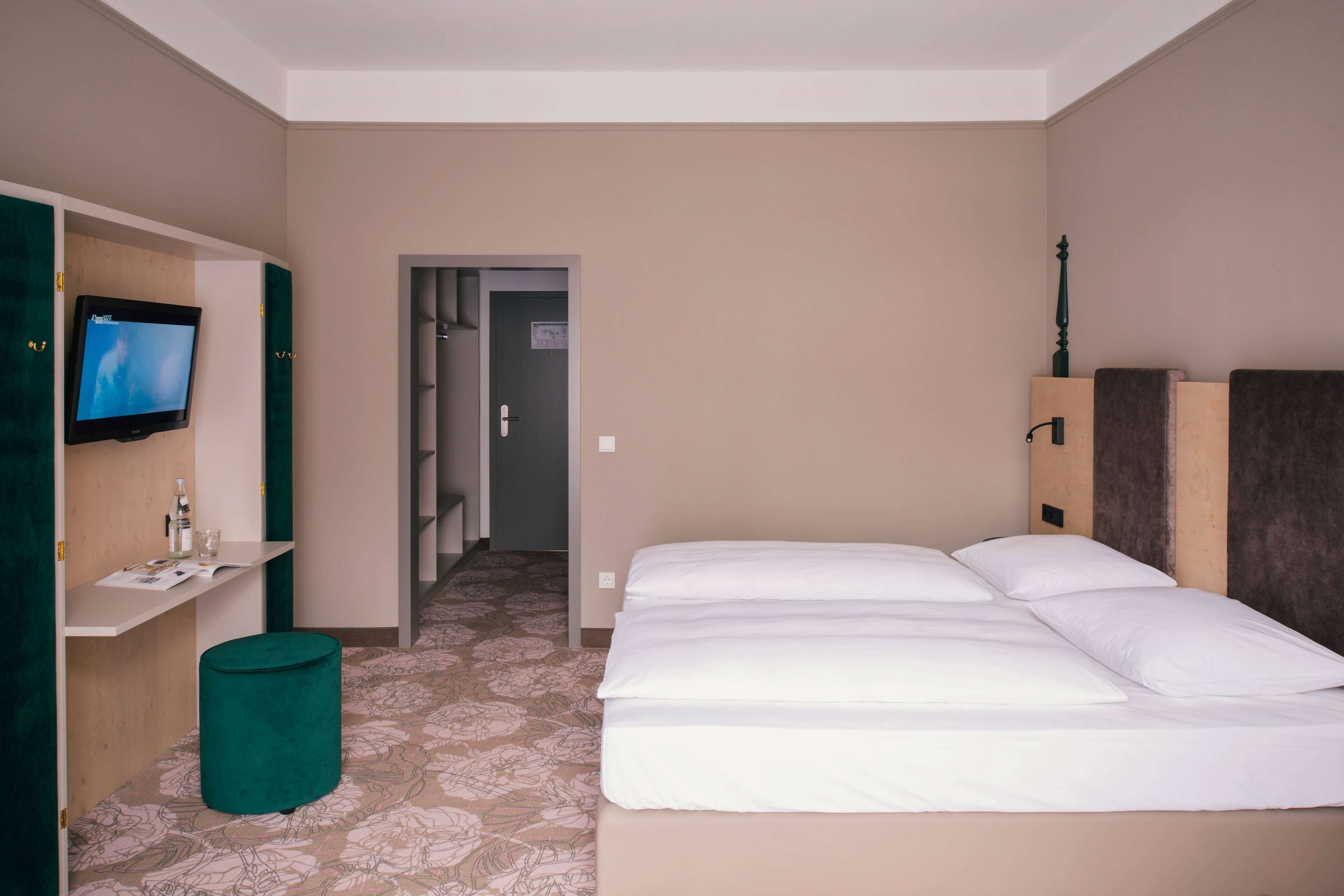 A hotel room with a bed, a television on a wall, a small table with a bottle and glass, and a trash bin.