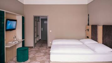 A hotel room with a bed, a television on a wall, a small table with a bottle and glass, and a trash bin.