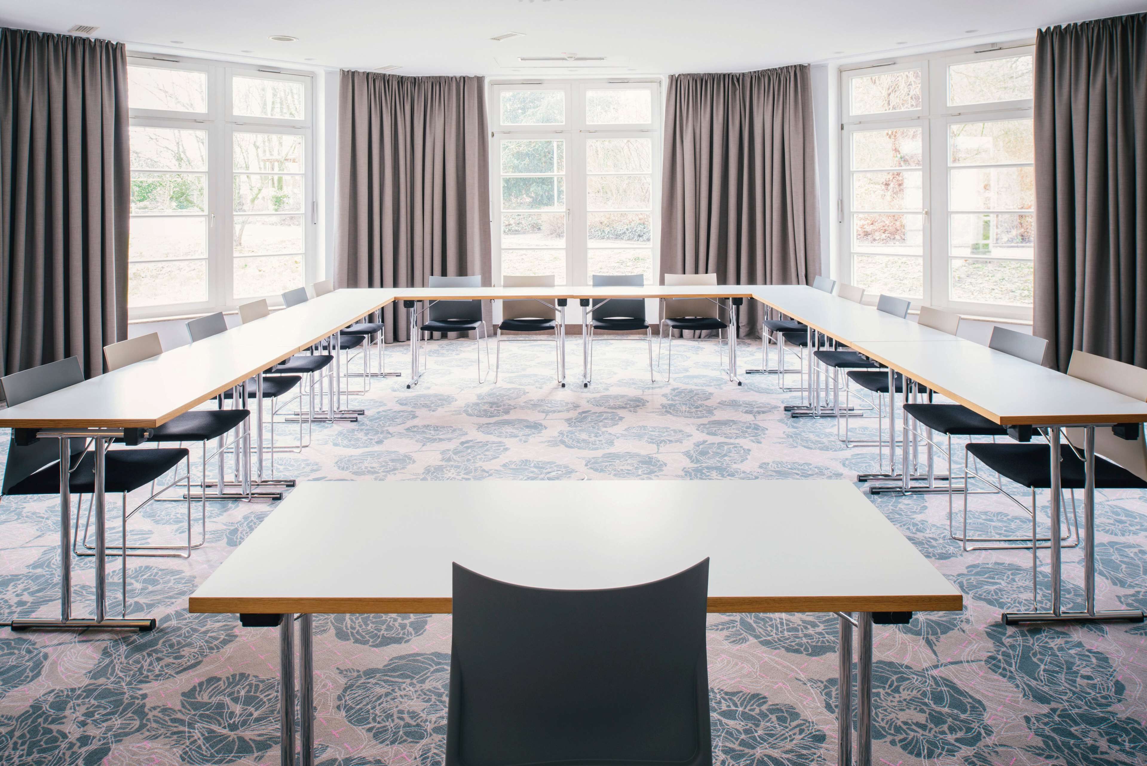 Uma sala de conferências com uma mesa comprida, várias cadeiras e janelas amplas com cortinas.