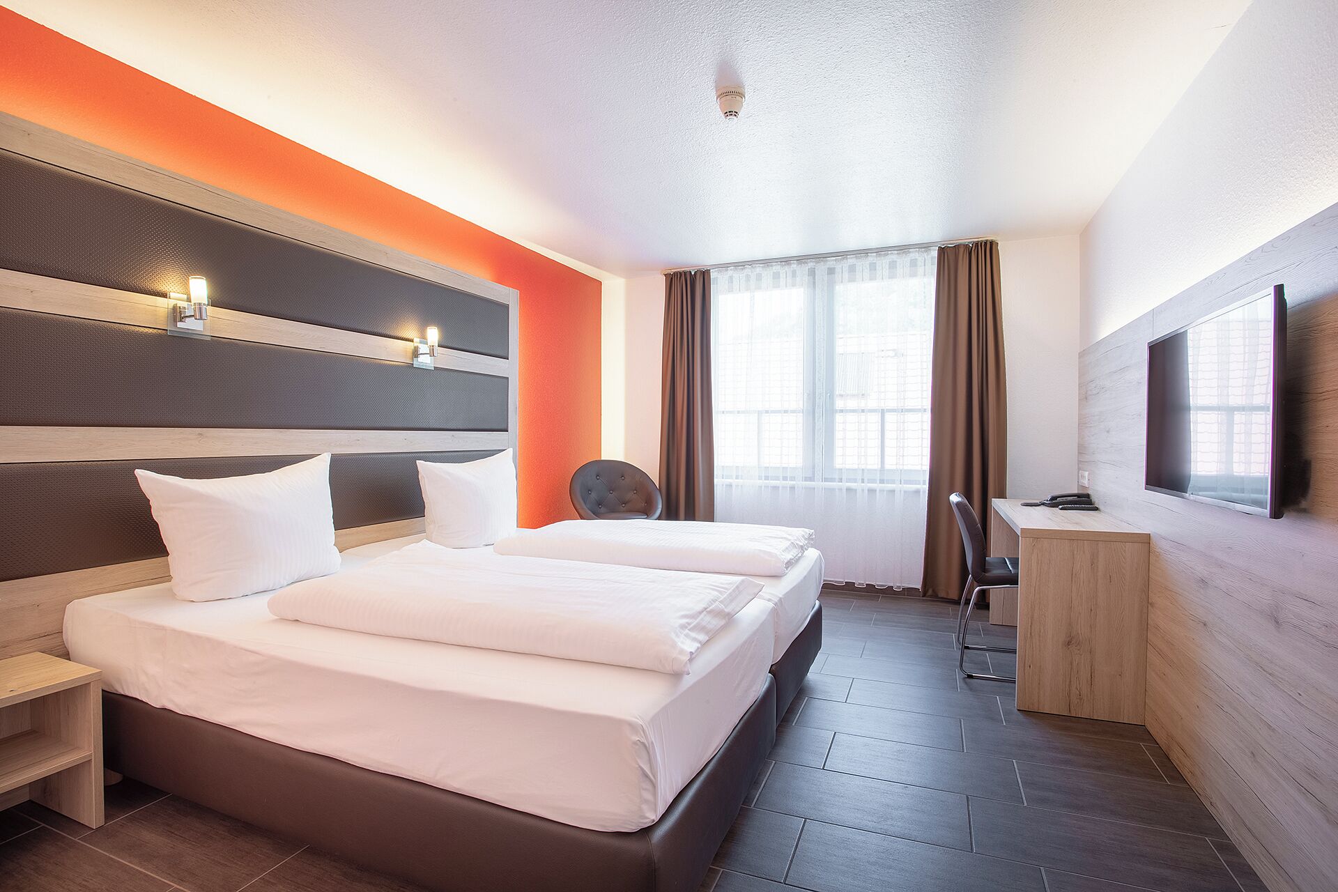 A hotel room with a double bed, a desk, a chair, a TV, and a window with curtains.