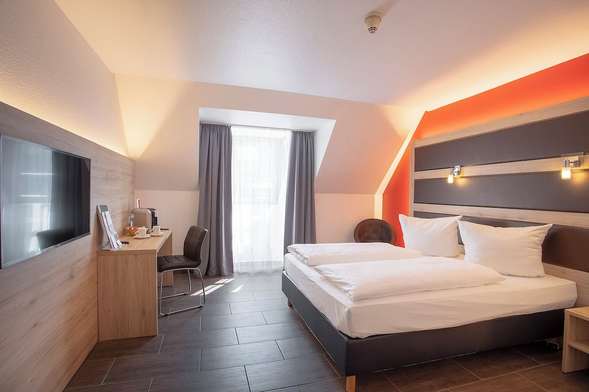 A hotel room with a bed, a desk with a chair, a TV, and a window with curtains.