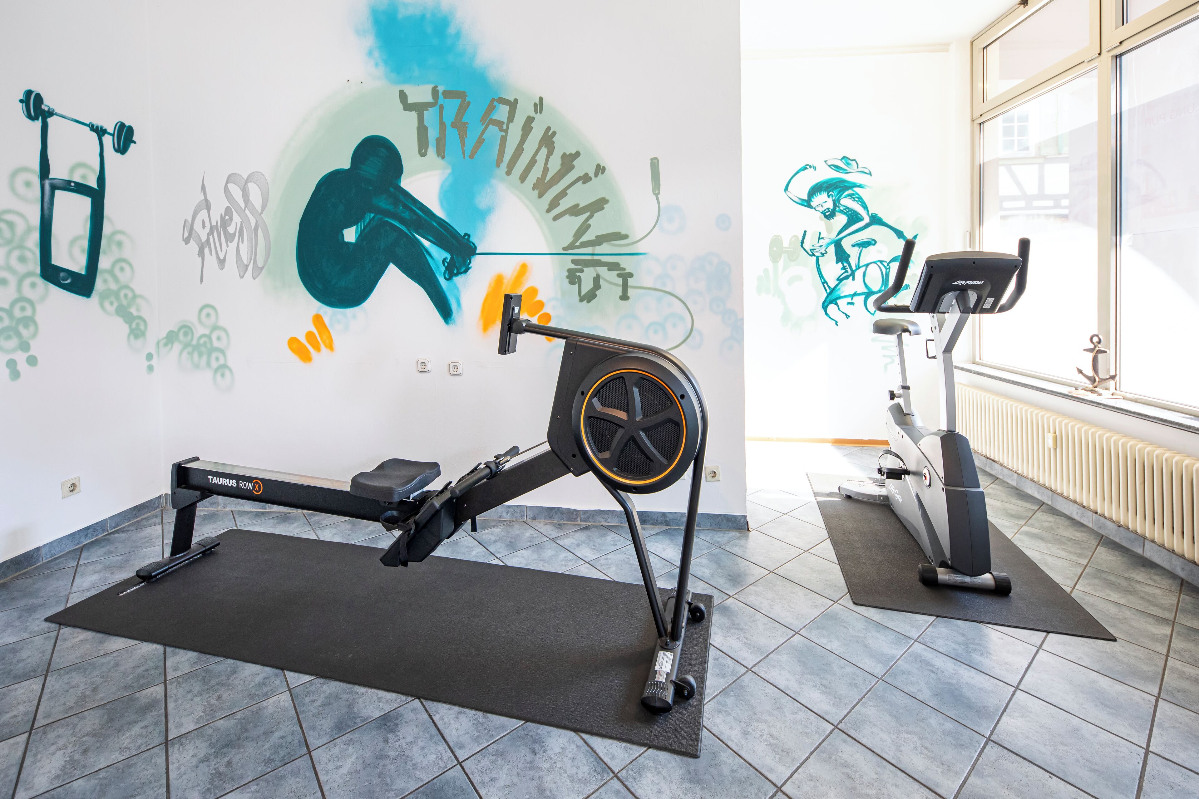 A gym with rowing machines and exercise bikes, a large window, and motivational wall art.