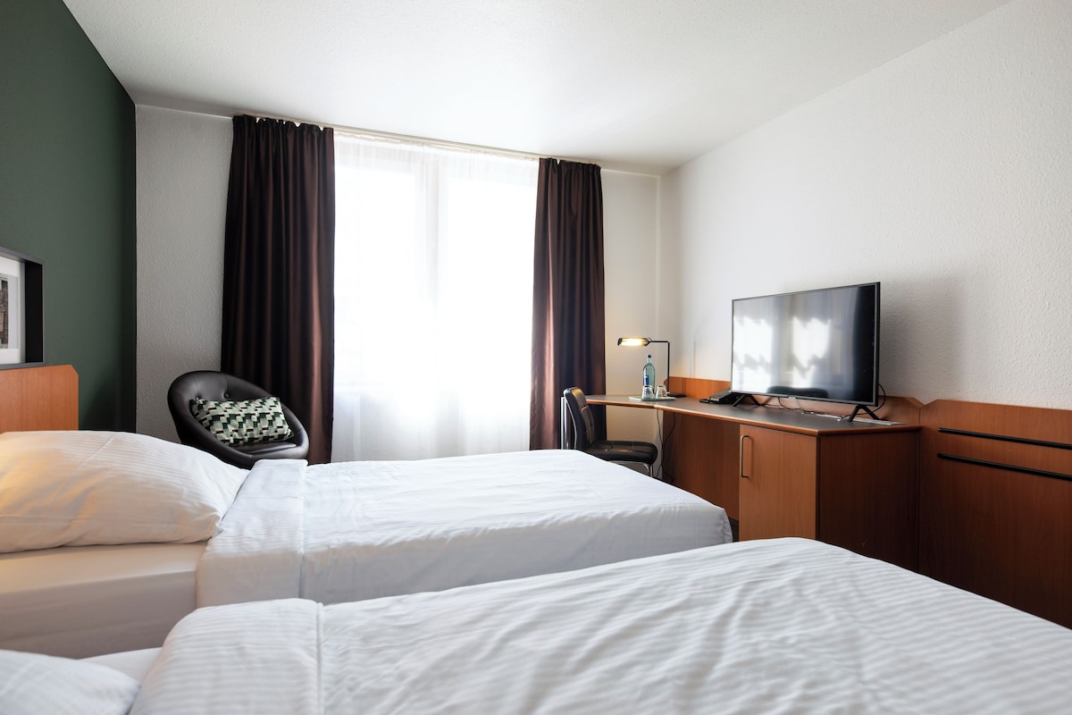 Una habitación de hotel con dos camas, un escritorio, una silla, un televisor y una ventana con cortinas.
