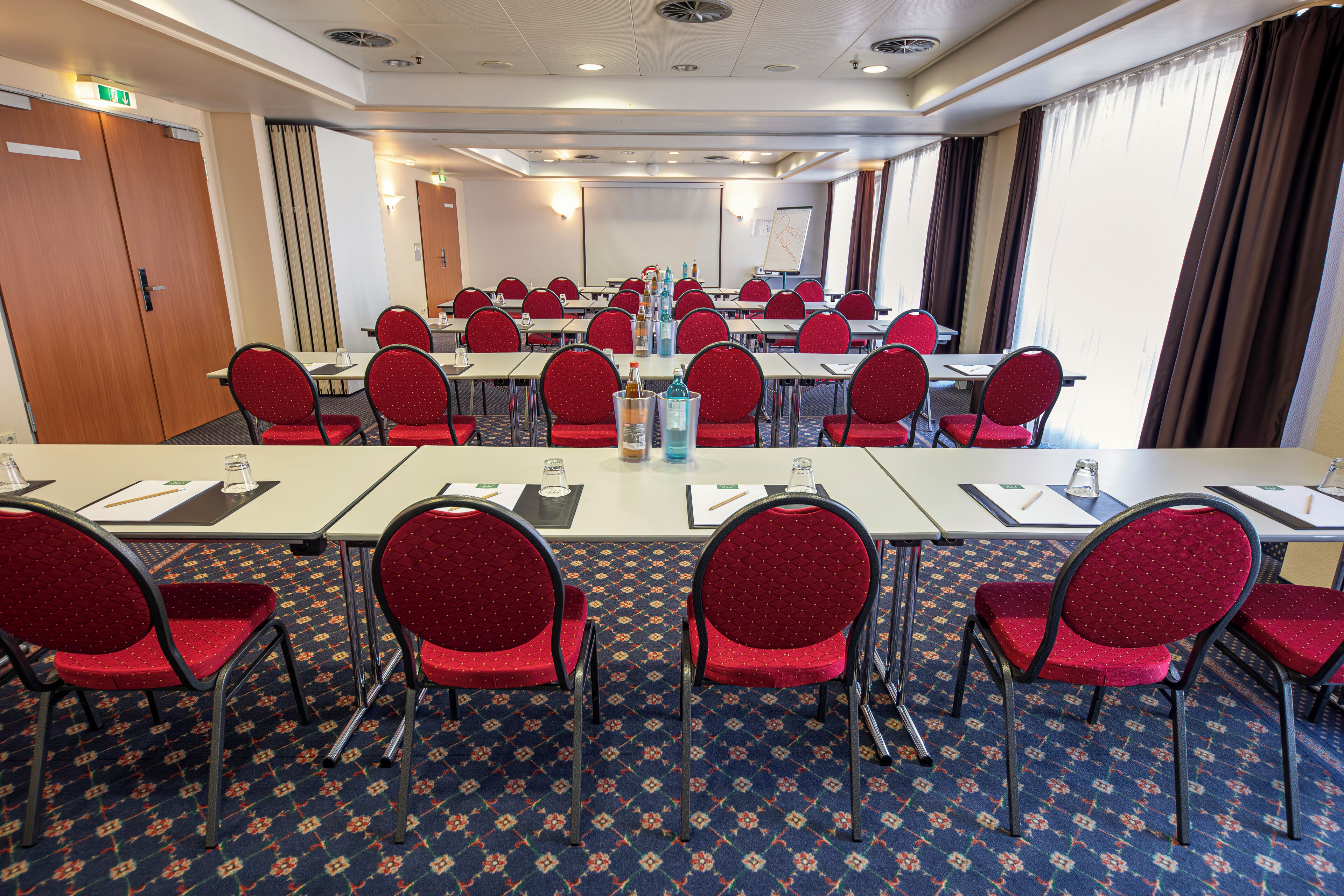 Une salle de conférence avec des rangées de tables et des chaises rouges, équipée de bouteilles d’eau et de blocs-notes.