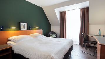 Ein Hotelzimmer mit einem großen Bett, einem Schreibtisch mit Stuhl, einem Bild an der Wand und einem Fenster mit Vorhängen.