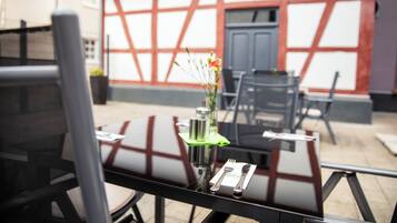 Un área de comedor al aire libre con una mesa para dos, un florero con flores y vistas a un edificio de estilo arquitectónico distintivo.