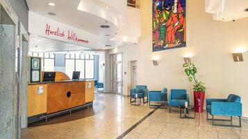 Zona de recepción de un hotel con un mostrador de madera, asientos azules, un cuadro grande y colorido y un letrero que dice "Herzlich Willkommen".