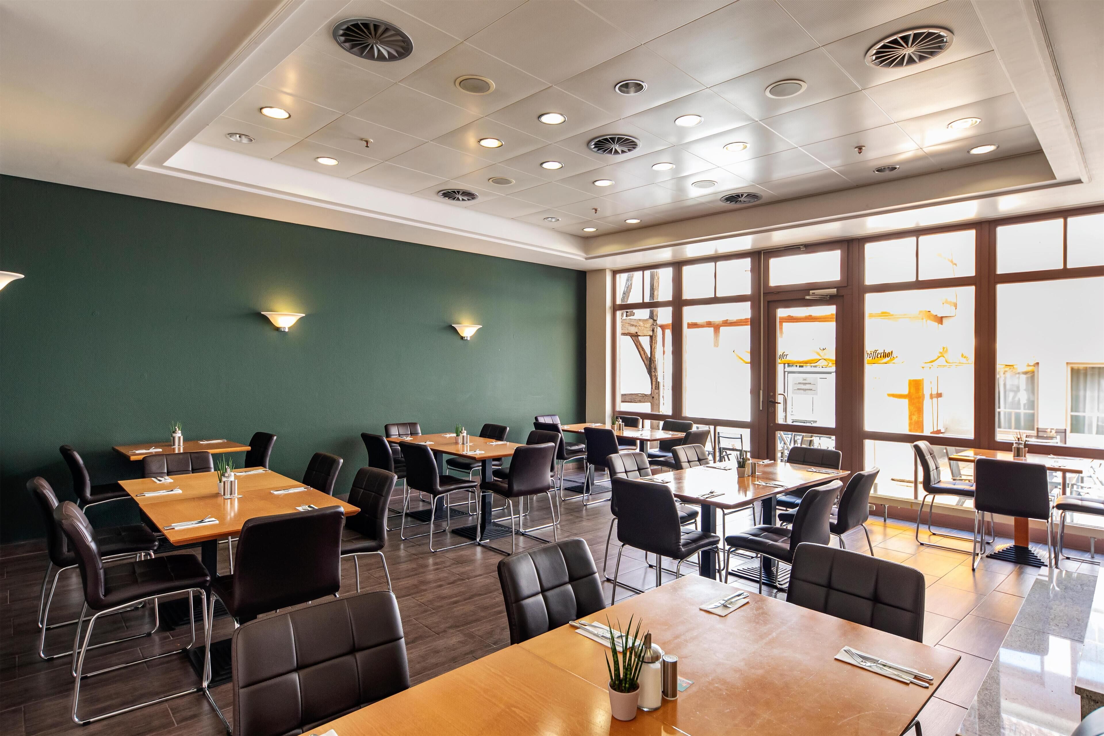 A restaurant with multiple tables and chairs, a green wall, and large windows allowing natural light.