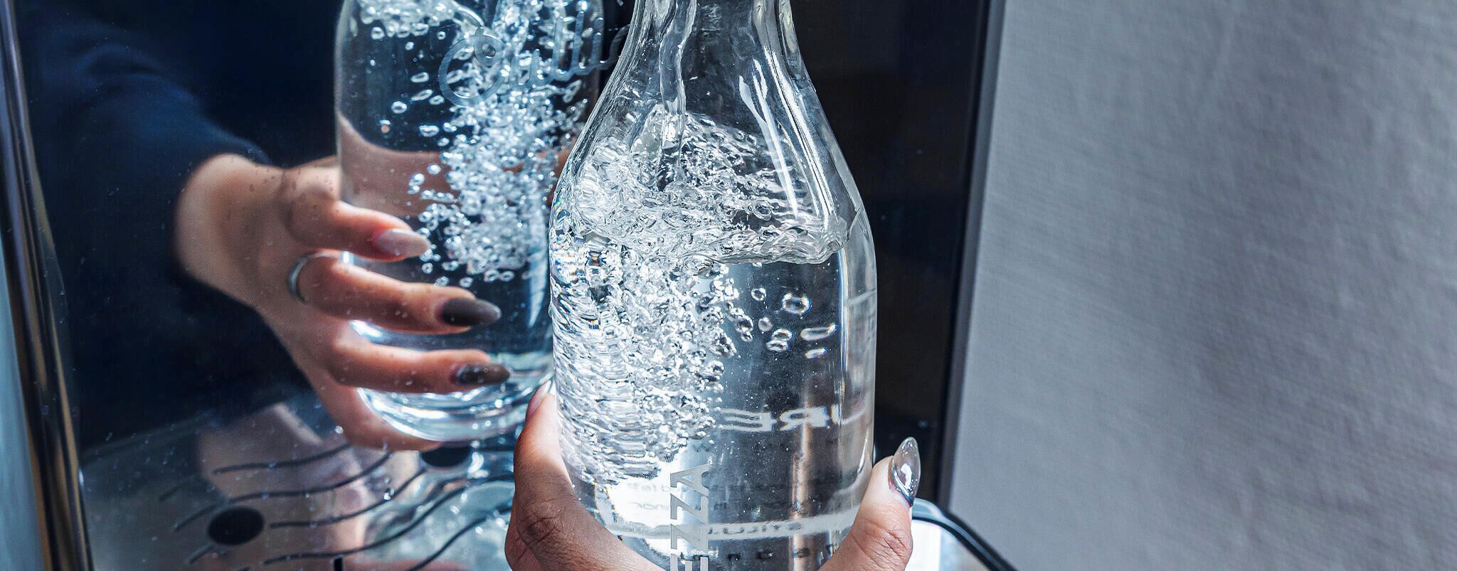 A person holding a clear glass bottle with visible bubbles, likely containing a carbonated beverage, with a reflection of a window in the background.