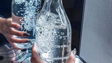 A person holding a clear glass bottle with visible bubbles, likely containing a carbonated beverage, with a reflection of a window in the background.