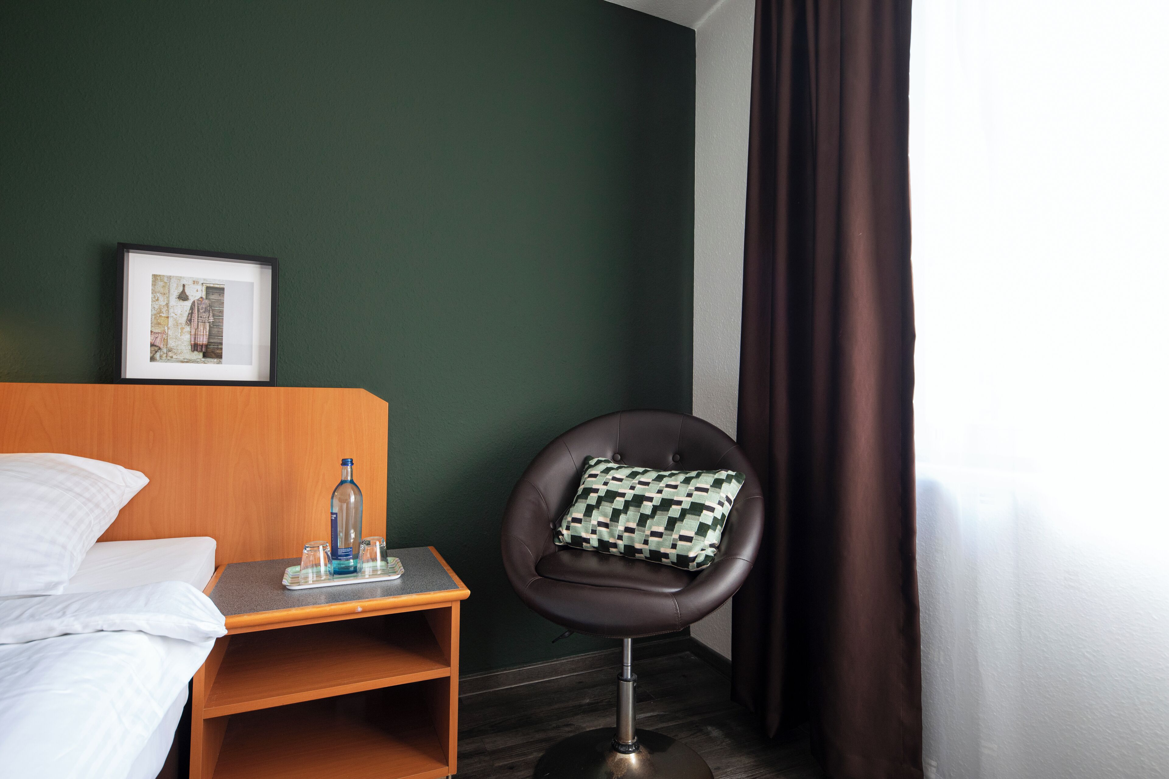 Una habitación de hotel con una cama, una mesita con una botella y vasos, una silla con un cojín estampado y un cuadro en la pared.
