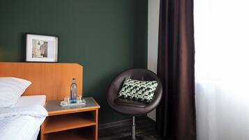 Una habitación de hotel con una cama, una mesita con una botella y vasos, una silla con un cojín estampado y un cuadro en la pared.