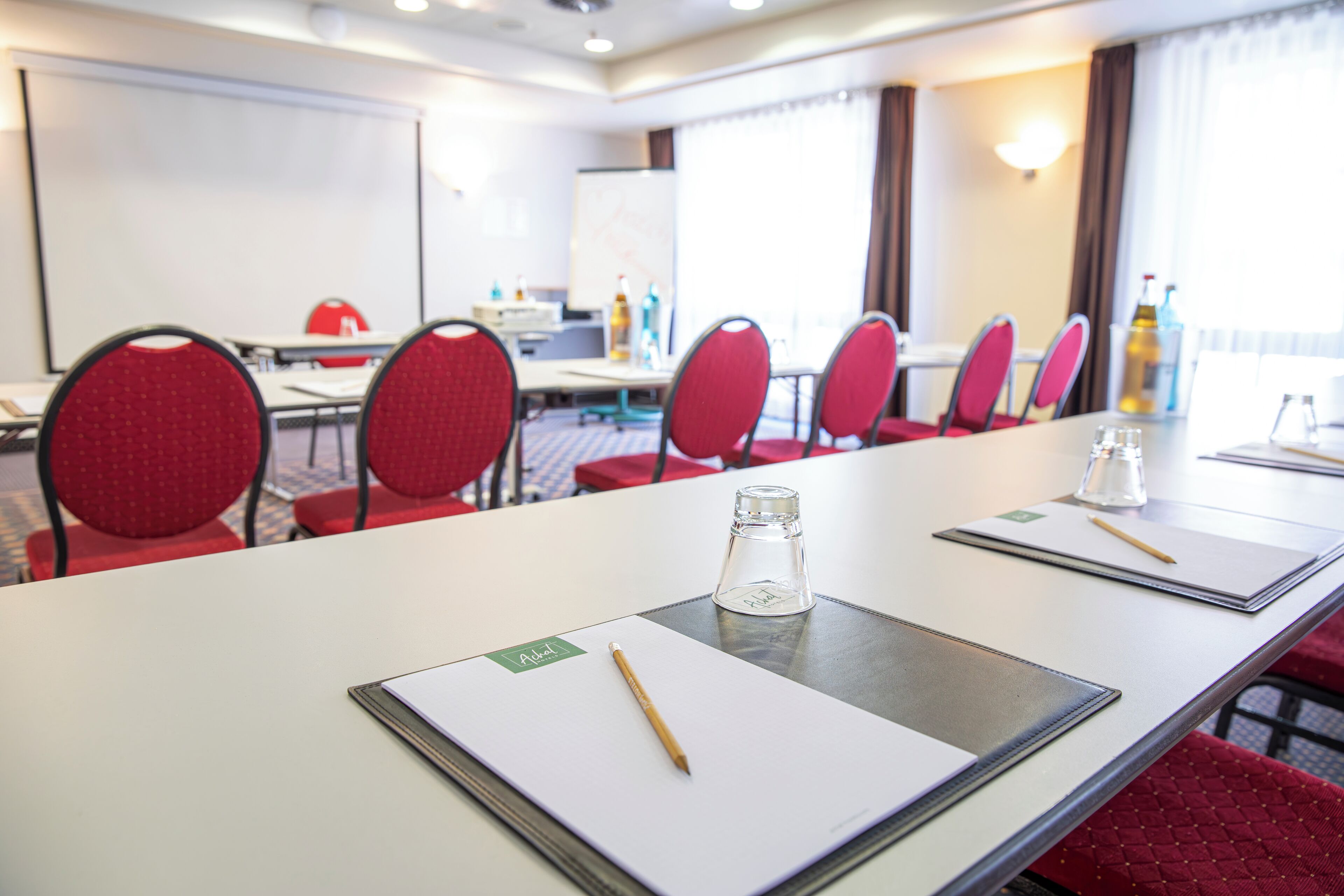 Une salle de conférence avec une longue table, des chaises rouges, un écran de projection et un tableau blanc.