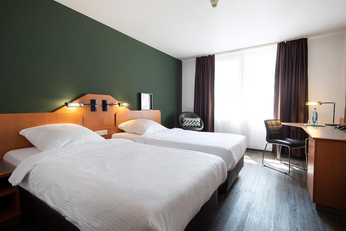 A hotel room with two beds, a desk, a chair, and a large window with curtains.
