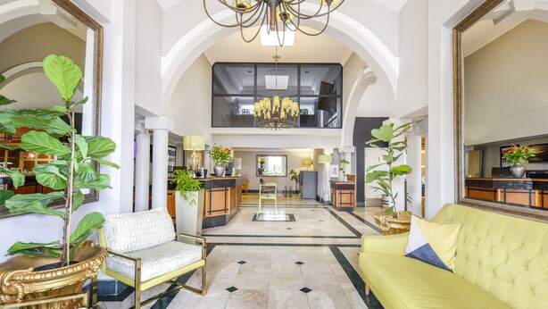 A spacious lobby with a high ceiling, a chandelier, and a central seating area with a yellow sofa and a white chair.