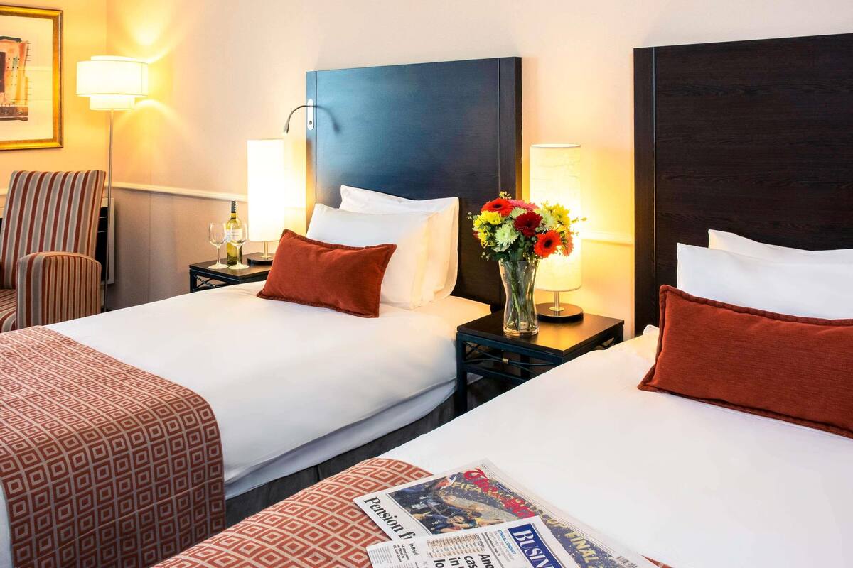 A hotel room with two beds, a nightstand with a lamp, a chair, and a vase with flowers.