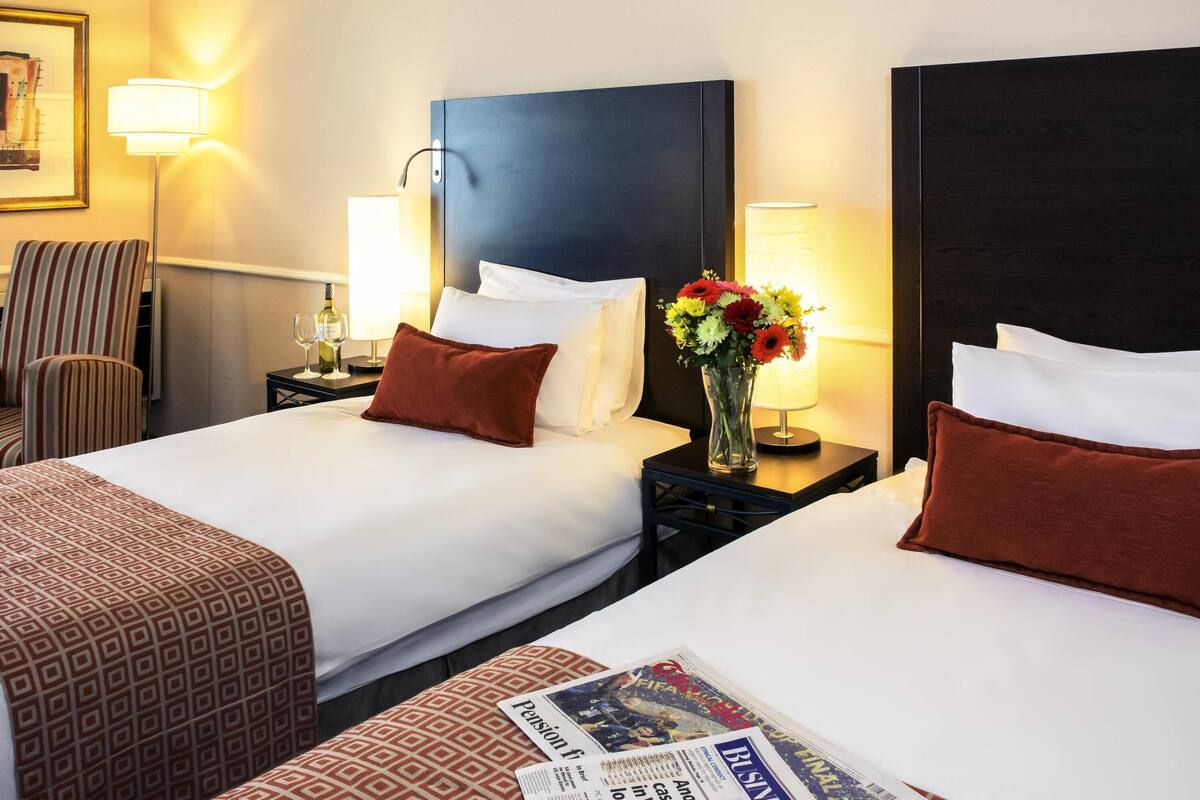 A hotel room with two beds, a nightstand with a lamp, a chair, and a vase with flowers.