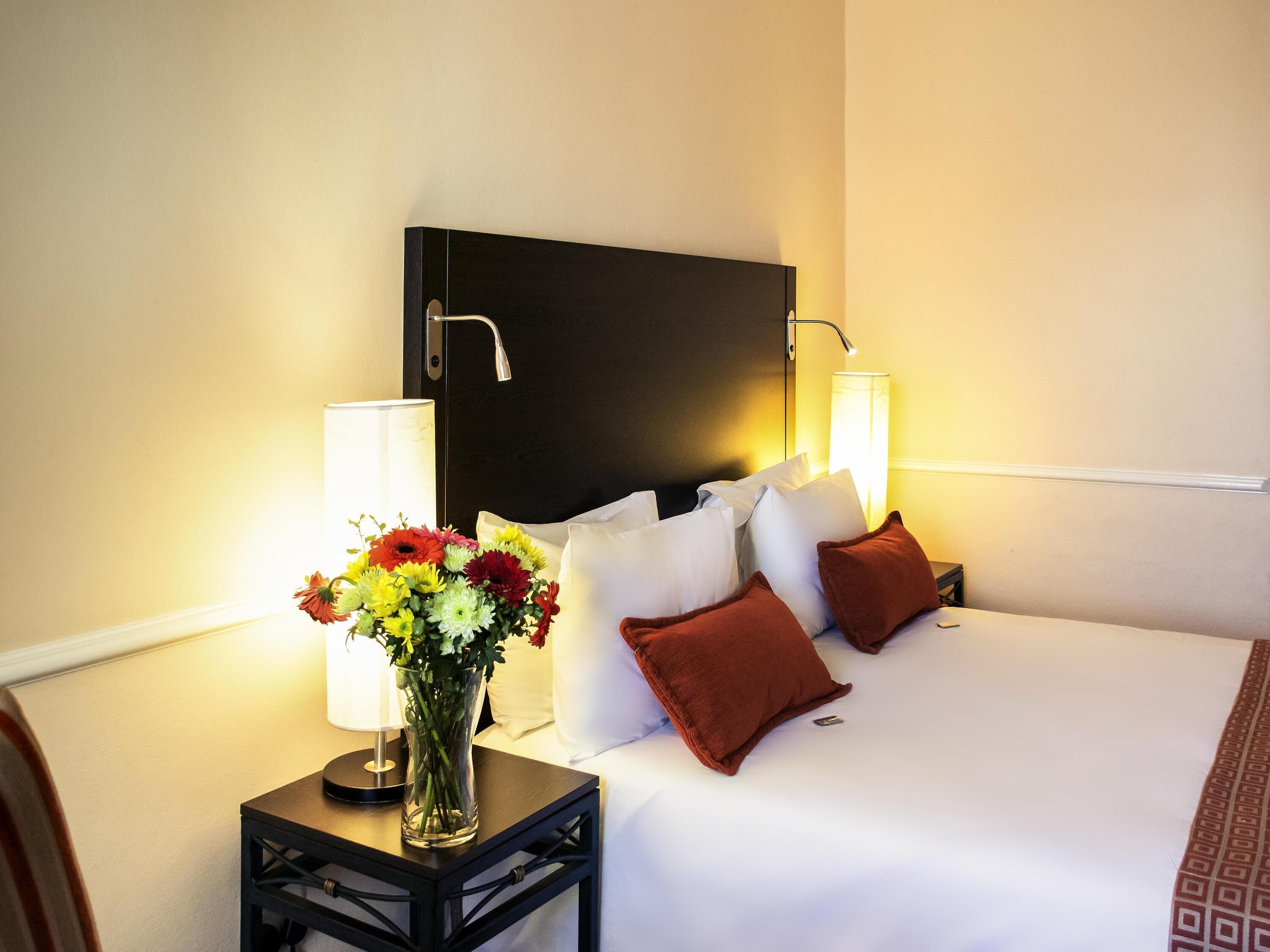 Quarto de hotel com cama, almofadas, um vaso com flores, uma mesa de cabeceira e uma luminária na parede.